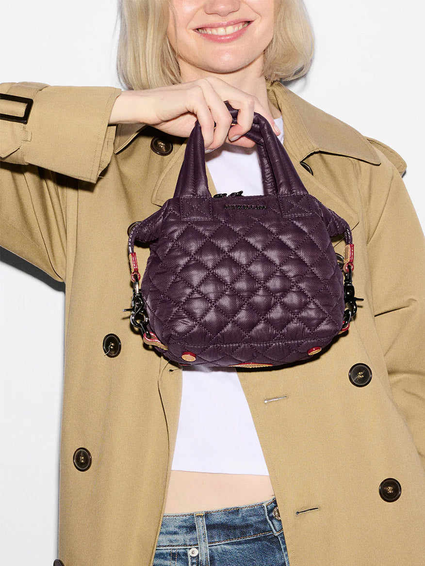 Wearing a beige trench coat and jeans, a person smiles at the camera while holding the MZ Wallace Micro Sutton in Raisin Oxford, a compact crossbody purse made from recycled nylon.
