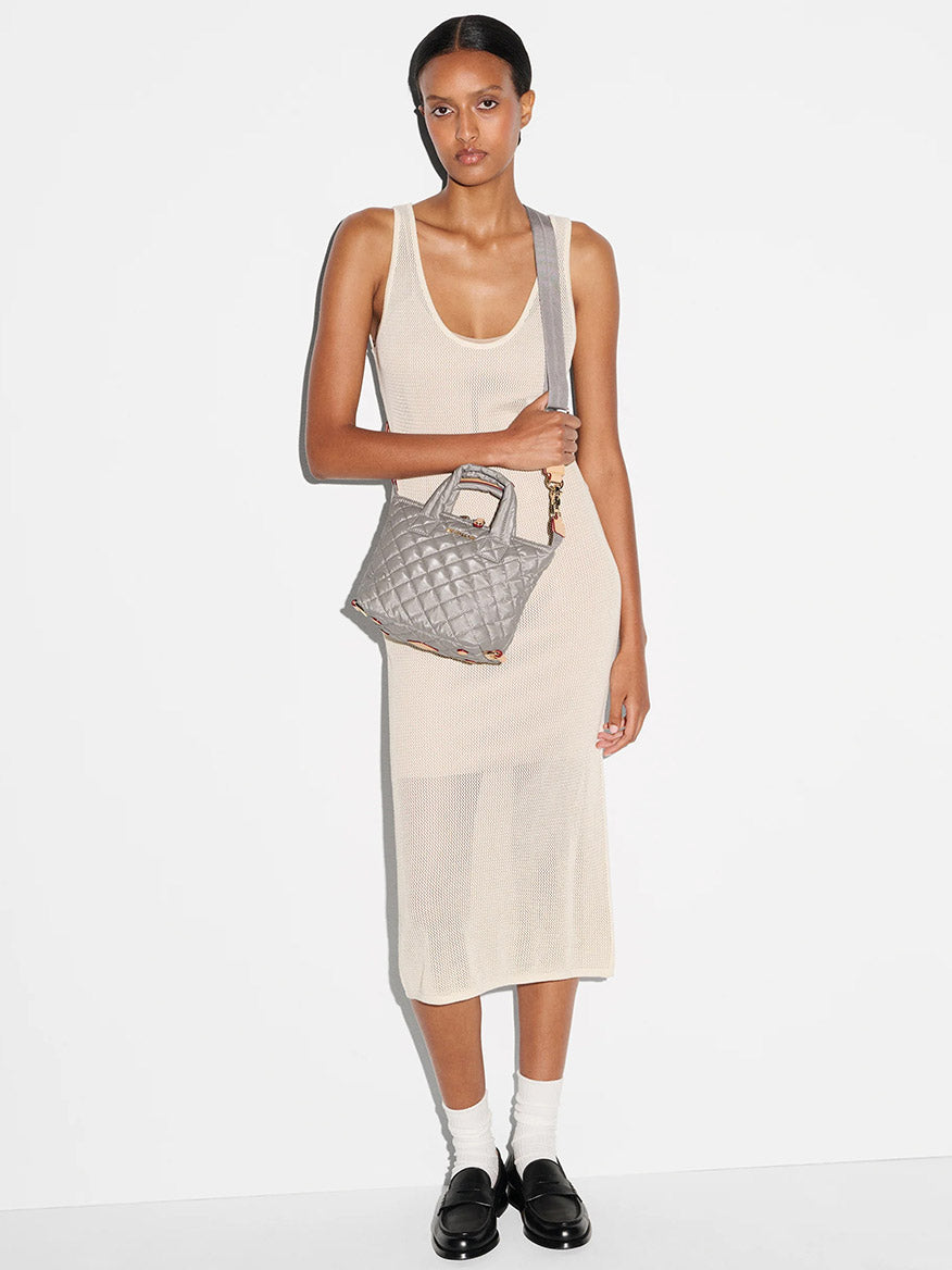 A woman in a sleeveless beige midi dress, white socks, and black loafers stands against a plain white background, holding the MZ Wallace Micro Sutton in Sable Pearl Metallic Oxford with an adjustable crossbody strap.