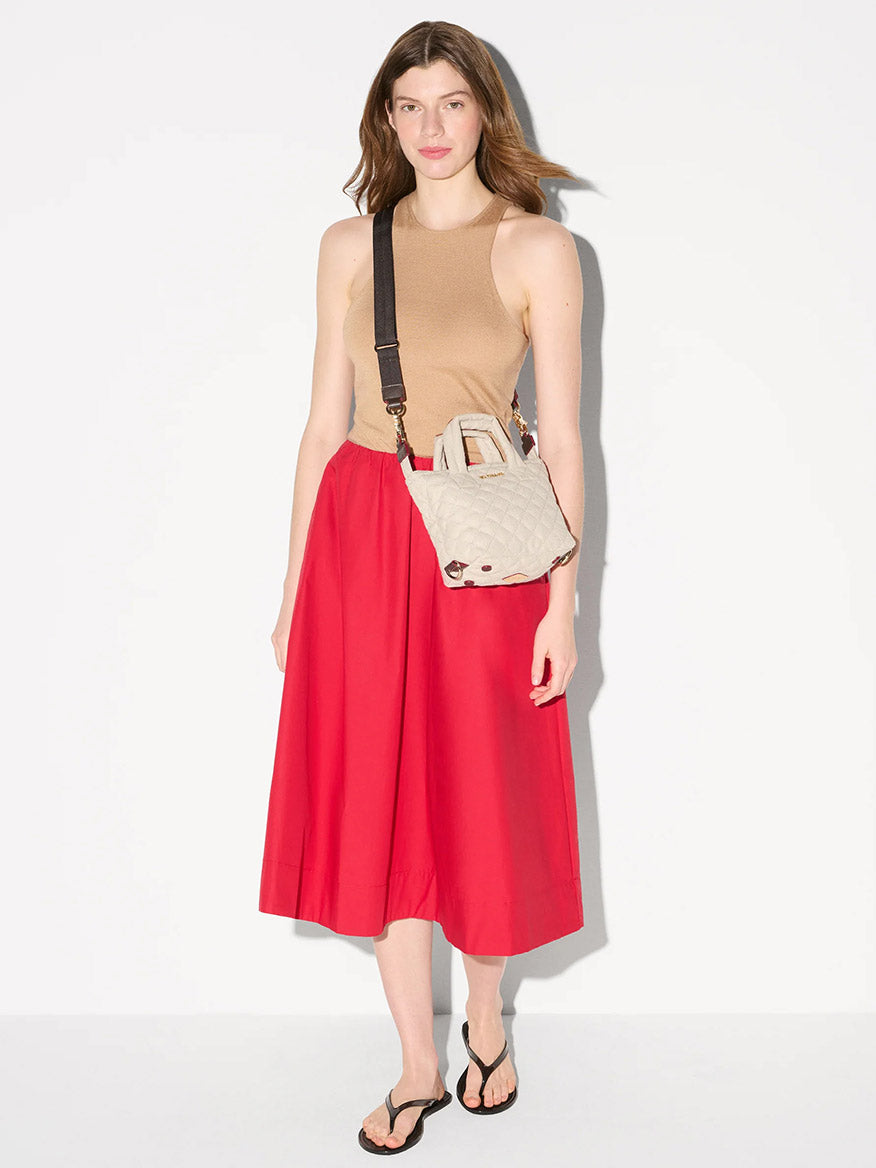 A woman stands against a plain background, wearing a beige sleeveless top, red midi skirt, black sandals, and carrying the MZ Wallace Micro Sutton in Sandy Linen Fabric for a structured carry.