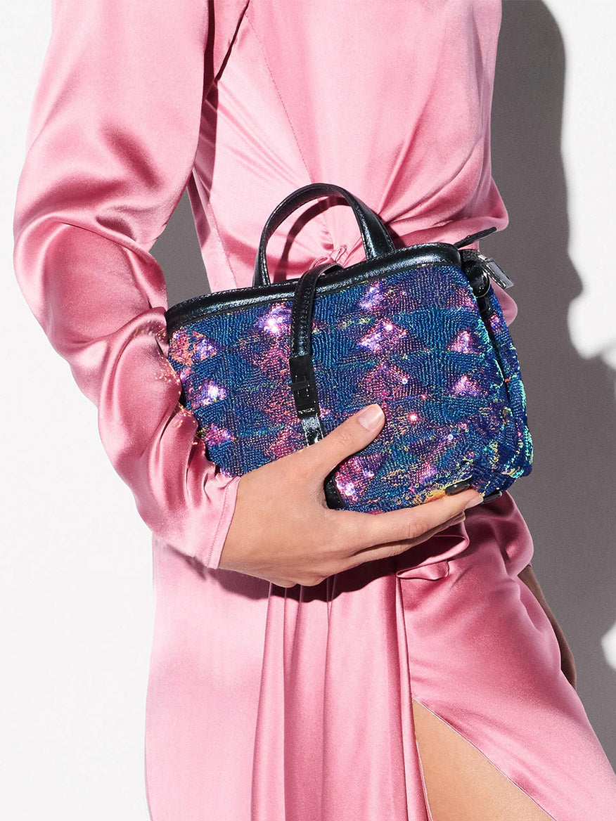 A person in a pink satin dress holds the MZ Wallace Mini Astor Top Handle in Aurora Sequin, a small blue and purple sequin handbag from the Astor Collection, featuring black handles and a buckle detail.
