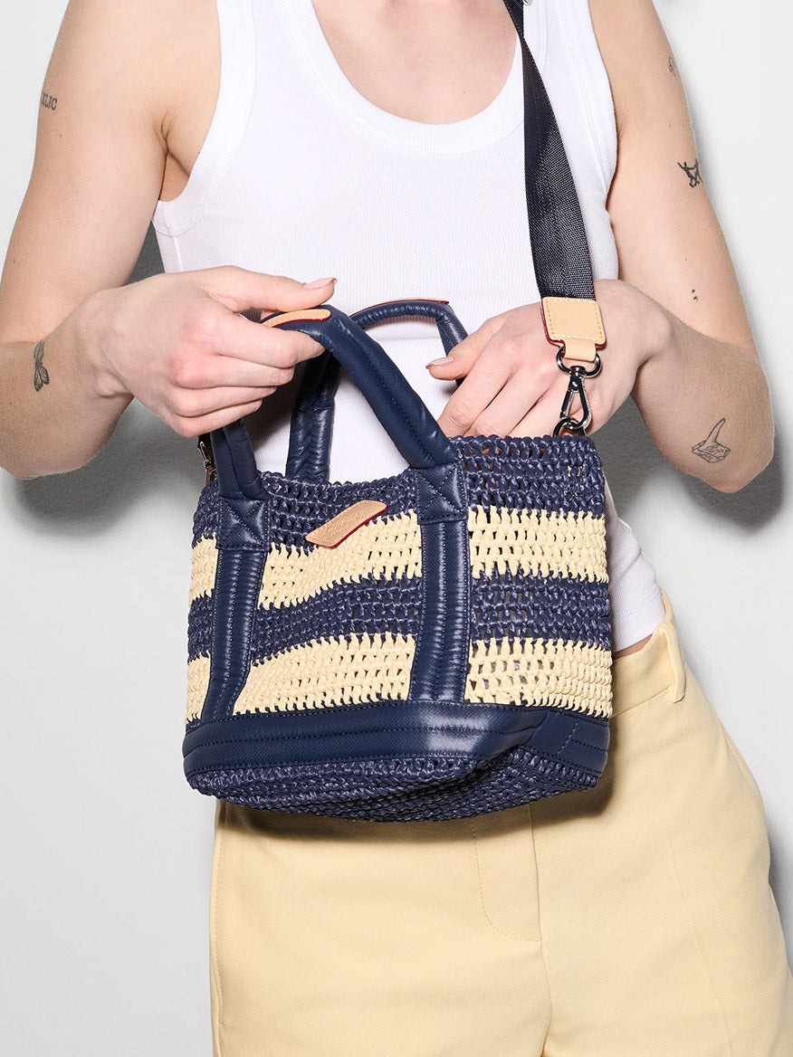 A person in a white tank top and beige pants holds the MZ Wallace Mini Crochet Tote in Dawn/Natural Stripe Crochet, a small blue and beige bag with a black shoulder strap.