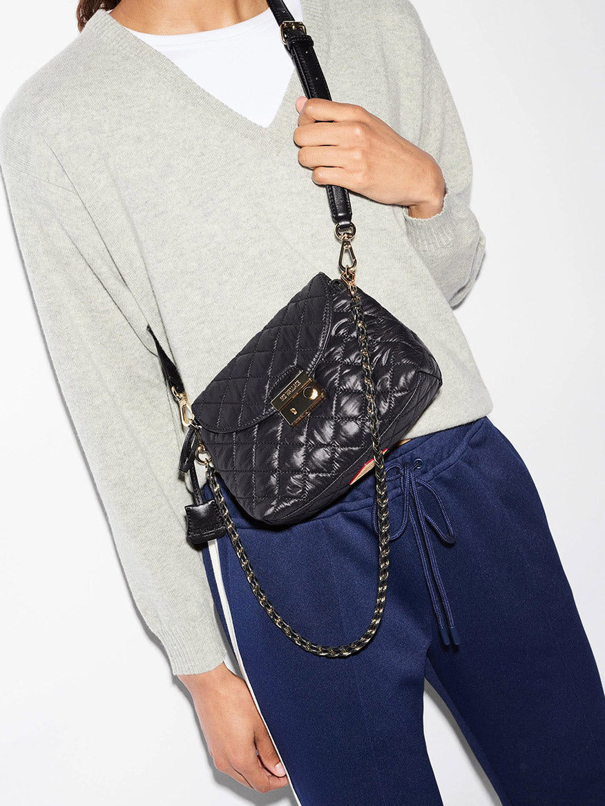 A person wears an MZ Wallace Mini Crosby Lock Shoulder in black, a small quilted recycled oxford nylon crossbody bag with a gold metal clasp and a chain-link strap.