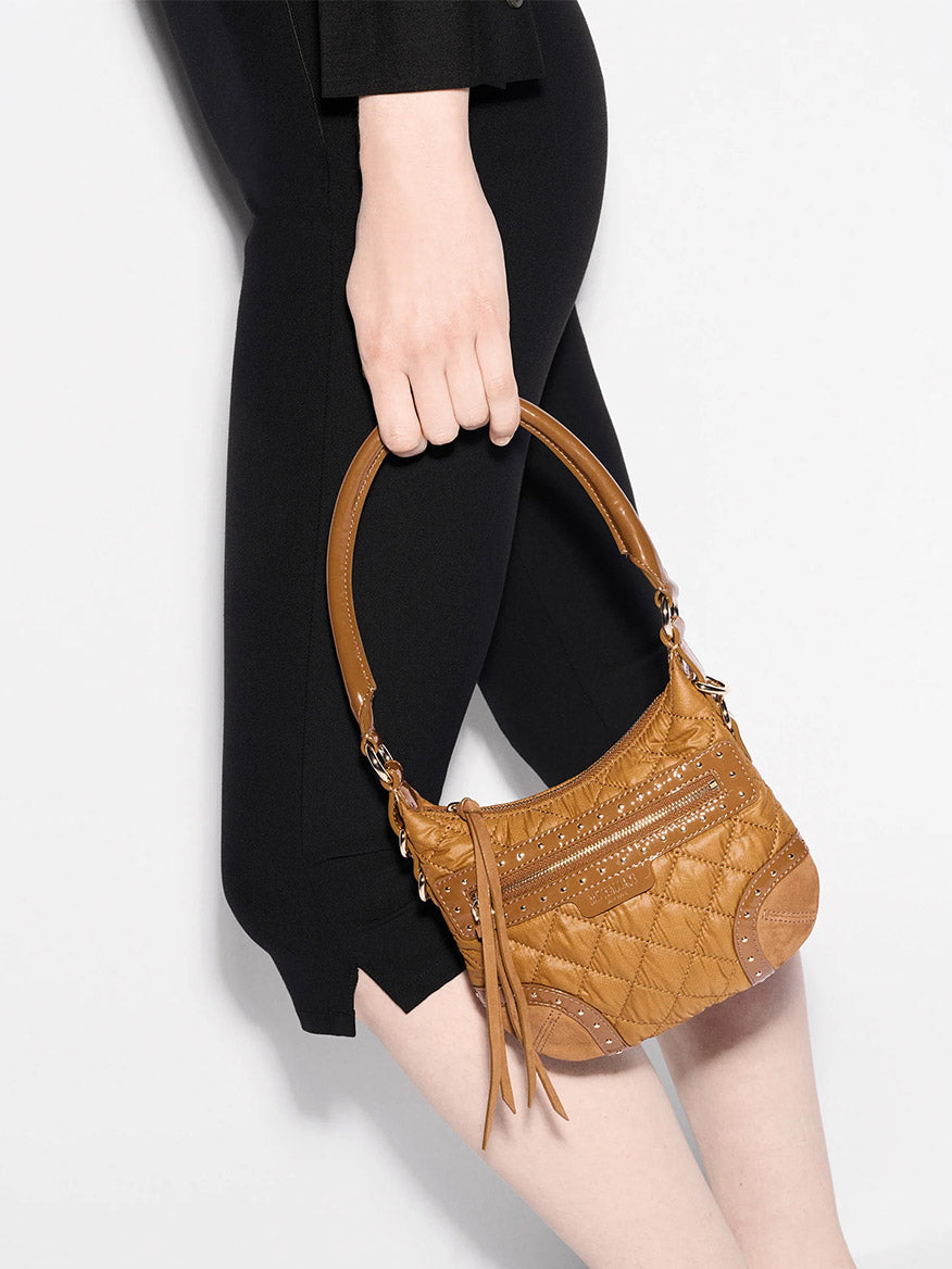 A person in a black outfit holds the MZ Wallace Mini Multi Stud Crosby Sienna in Amber Stud & Suede, a small brown quilted handbag with Italian leather trim, short handle, gold hardware, and decorative tassels.
