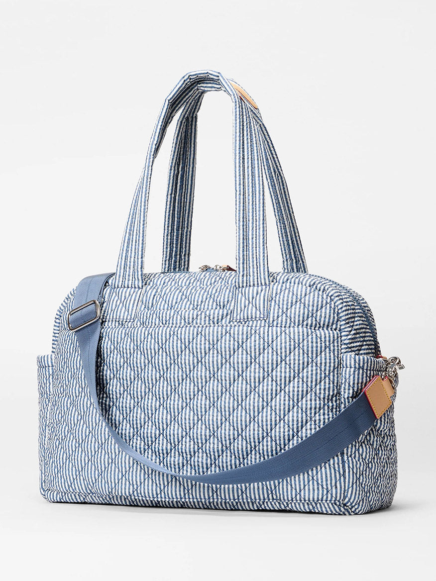 The MZ Wallace Nik Duffel in Seersucker Oxford, featuring a blue and white striped quilted pattern, two handles, a detachable shoulder strap, and zippered top closure, is shown against a plain white background—ideal for travel.