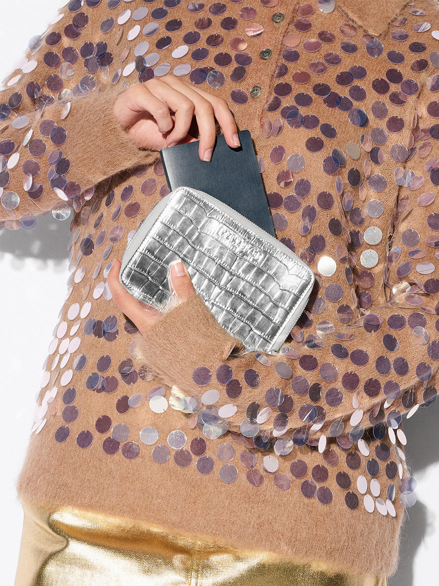 A person wearing a brown sequin sweater holds the MZ Wallace Passport Organizer in Silver Croc Effect and a black smartphone against a white background.