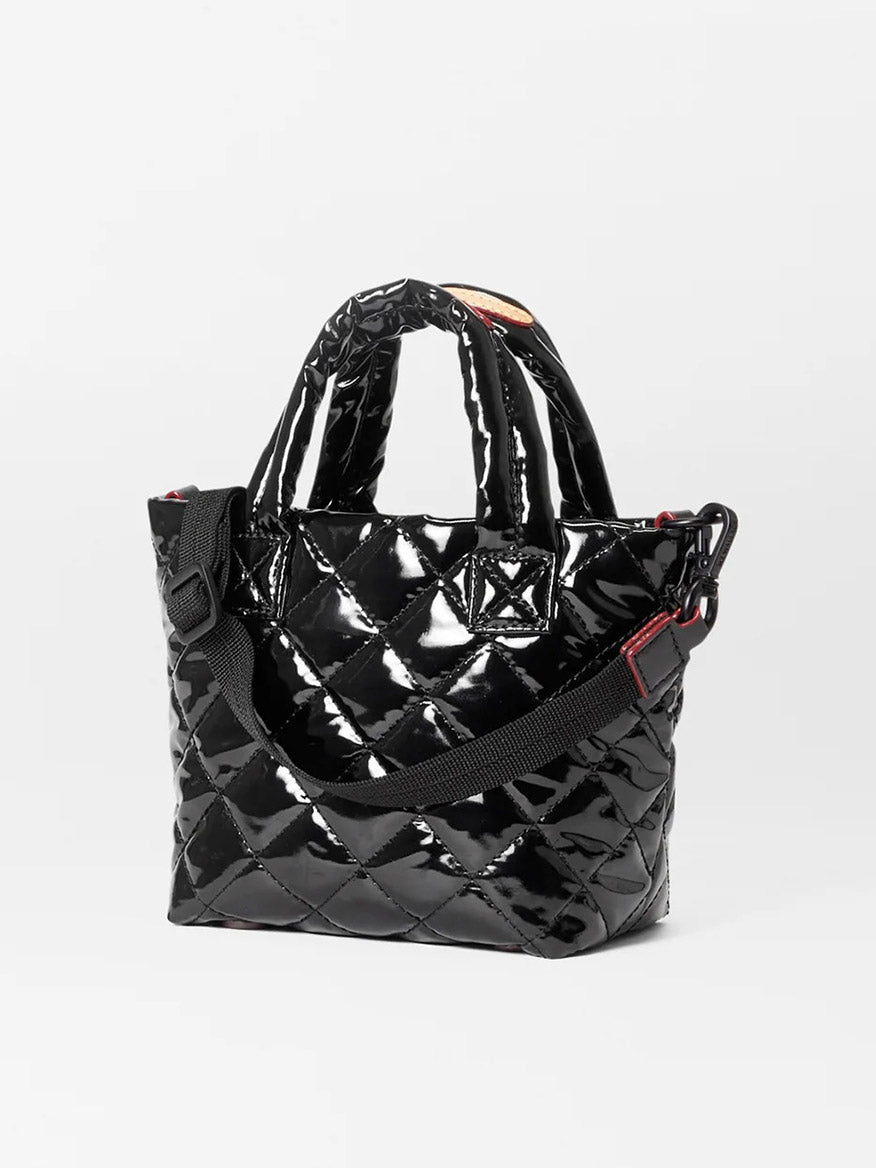 The MZ Wallace Petite Metro Tote Deluxe in Black Lacquer Oxford is a small, black quilted mini nylon bag with a glossy finish. It features short handles, an adjustable black shoulder strap, and Italian leather trim for an added touch of elegance.
