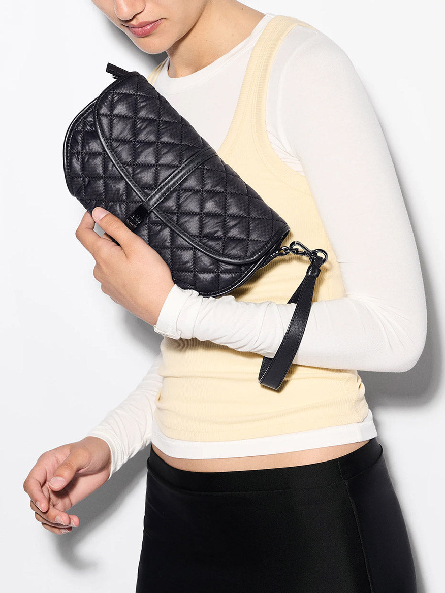 A person in a cream long-sleeve top under a yellow tank holds the MZ Wallace Small Astor Convertible Clutch in Black Oxford.