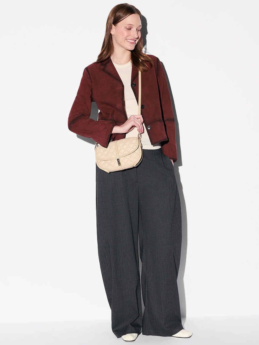 A person stands against a plain white background, holding the MZ Wallace Small Astor Convertible Clutch in Buff Oxford; they wear a burgundy jacket, cream top, wide-leg gray trousers, and white shoes.