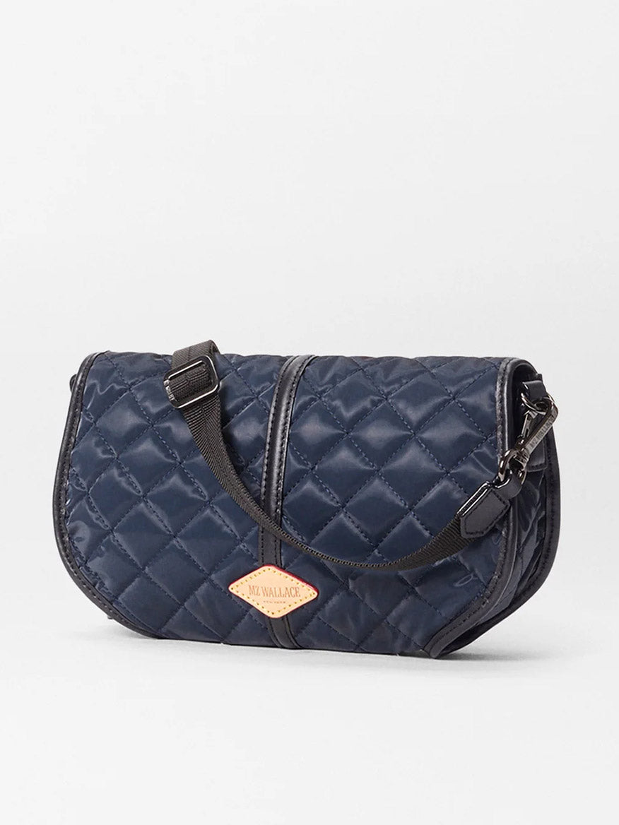 The MZ Wallace Small Astor Convertible Clutch in Ink Liquid Oxford features navy quilted fabric, a black adjustable strap, and a tan logo patch on the front, shown against a plain white background.