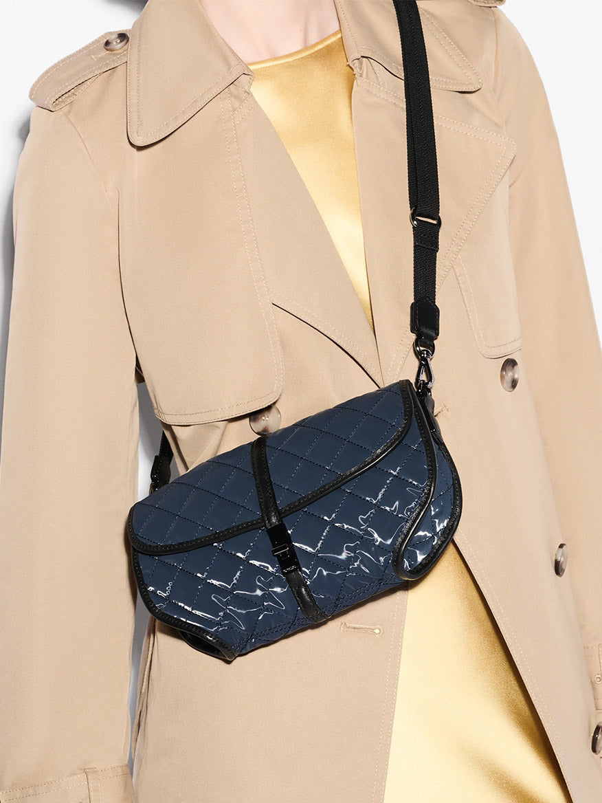 A person in a beige trench coat and gold top carries the MZ Wallace Small Astor Convertible Clutch in Ink Liquid Oxford, a quilted navy blue bag with an adjustable black strap.