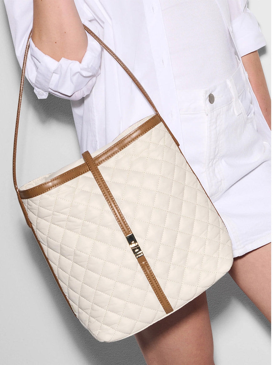 A person in white clothes holds the MZ Wallace Small Astor Hobo in Sandshell Oxford With Brown, a chic shoulder bag featuring quilted nylon, Italian leather trim, and a brown strap.