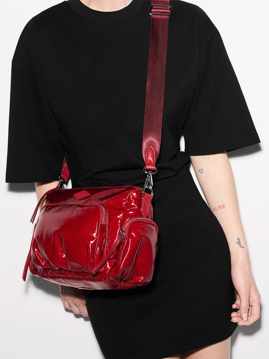 Person wears a black t-shirt dress and carries the MZ Wallace Small Chelsea Crossbody in Apple Gloss Bedford—a shiny, quilted nylon bag with multiple compartments and adjustable strap. Tattoos are visible on their forearm and wrist.