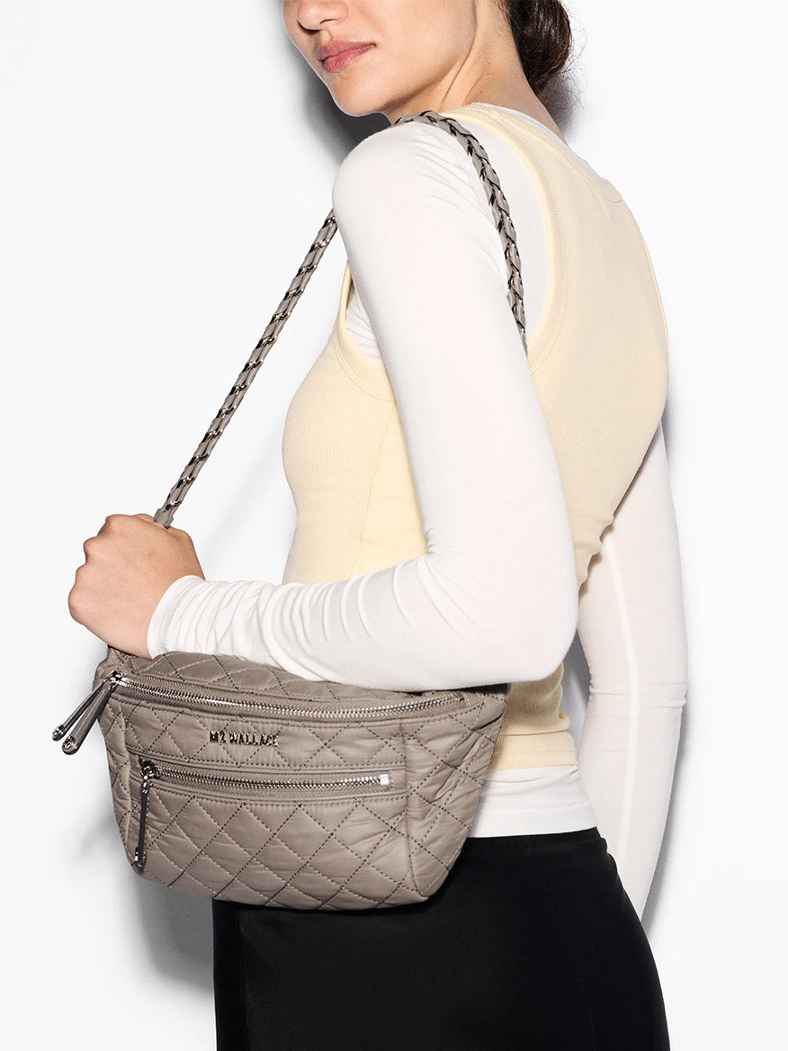 A person wearing a sleeveless beige top over a long-sleeve white shirt carries the MZ Wallace Small Crosby Sling Bag in Morning Dove Oxford, a crossbody bag with quilted fabric and zippered pockets.