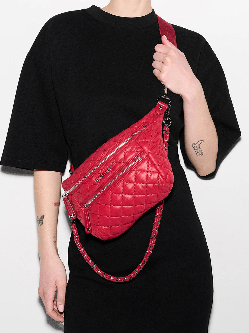 A person in a black dress poses with the MZ Wallace Small Crosby Sling Bag in Apple Oxford, a quilted red crossbody featuring silver zippers and a chain strap.