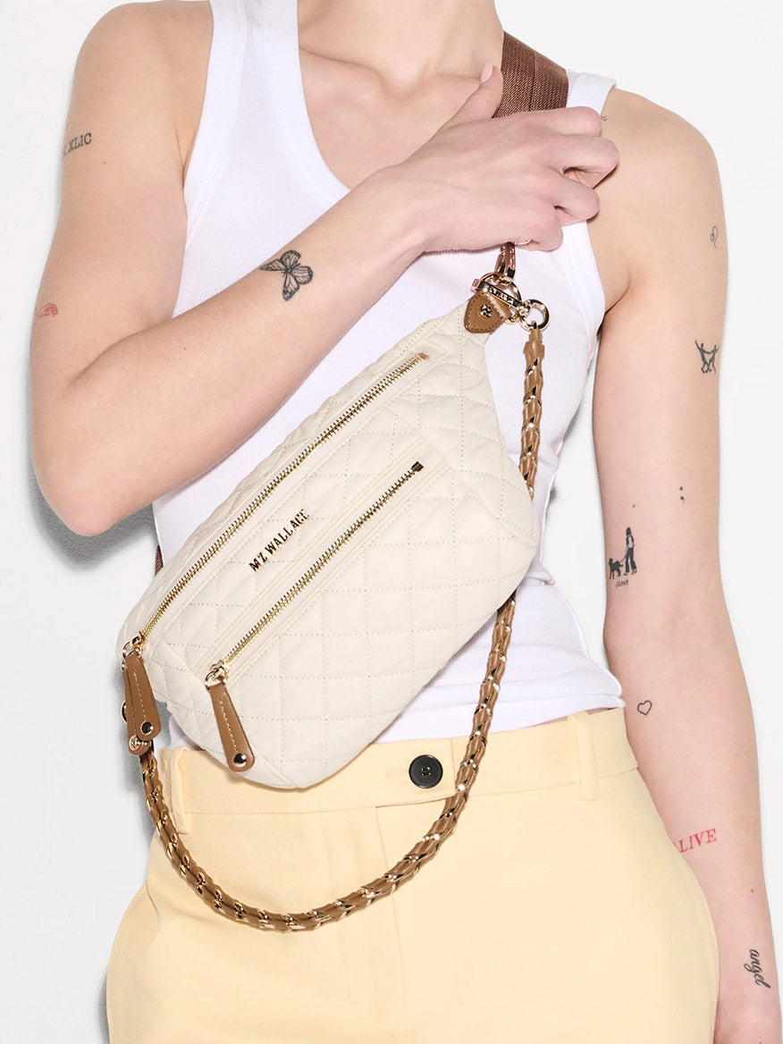 A person in a white tank top and yellow pants holds the MZ Wallace Small Crosby Sling Bag in Sandshell Oxford with Brown.
