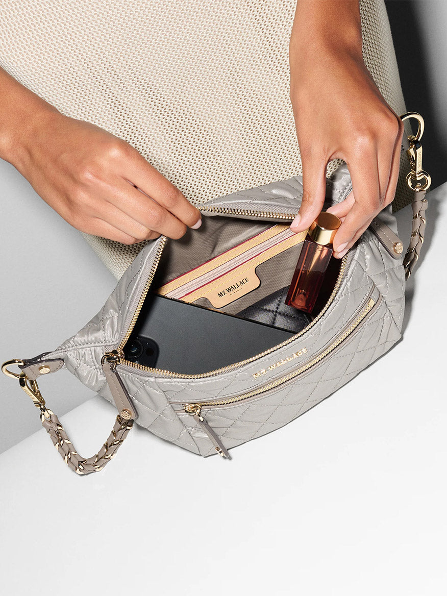 A person opens an MZ Wallace Small Crosby Sling Bag in Sable Pearl Metallic Oxford, revealing a beige wallet, a small bottle, and a smartphone inside.