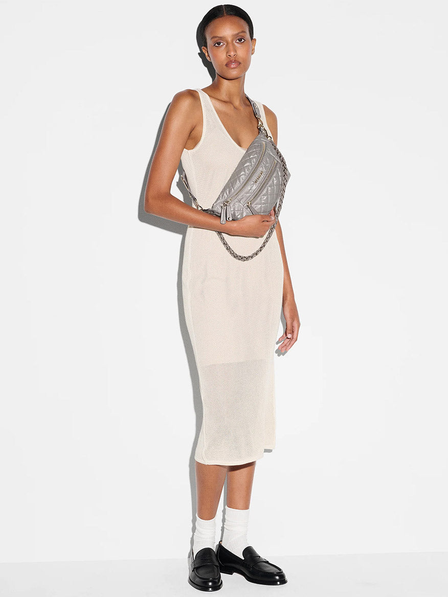 A person stands against a white background wearing a sleeveless beige dress, white socks, black loafers, and an MZ Wallace Small Crosby Sling Bag in Sable Pearl Metallic Oxford.