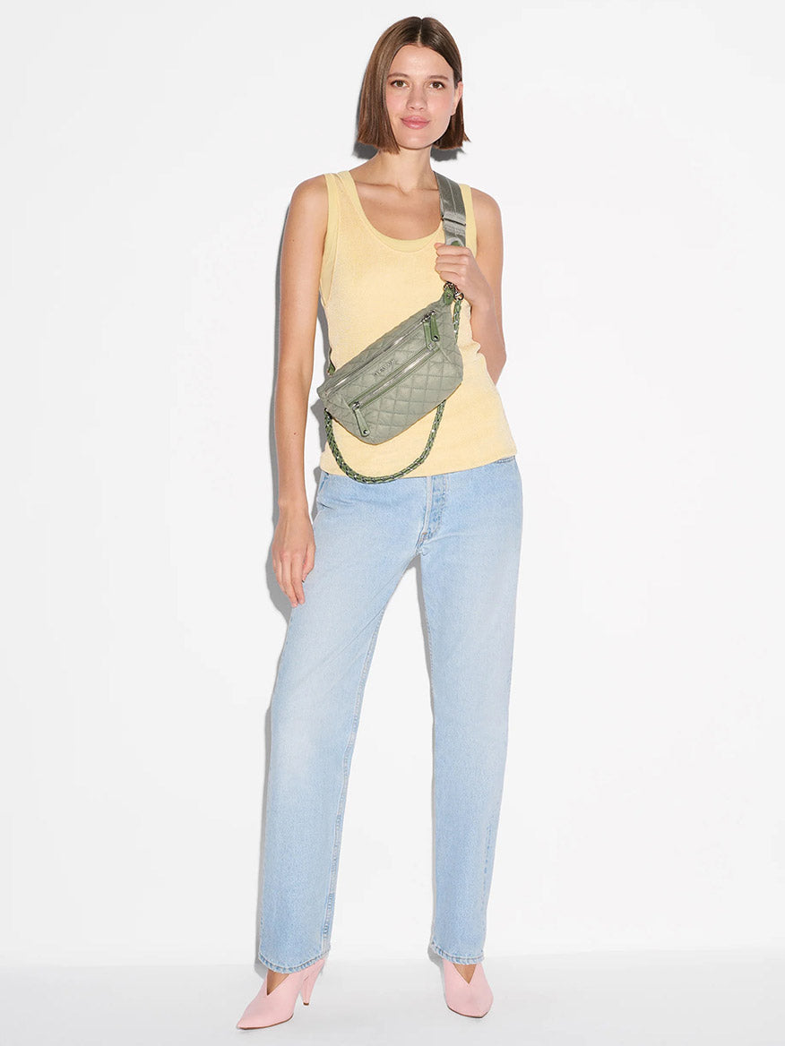 A person stands against a white background wearing a yellow tank top, light blue jeans, pink heels, and carrying the MZ Wallace Small Crosby Sling Bag in Agave Green Oxford with an adjustable strap.