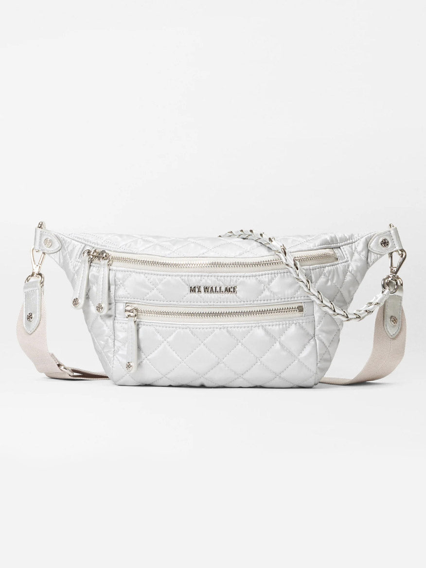 The MZ Wallace Small Crosby Sling Bag in Oyster Metallic Oxford features white quilted durable Oxford, silver zippers, a front pocket, and an adjustable beige strap, shown against a plain background.