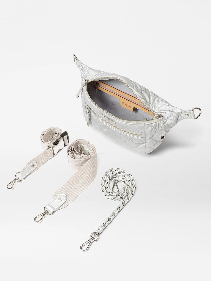 The MZ Wallace Small Crosby Sling Bag in Oyster Metallic Oxford is shown with a silver quilted finish, a partially open zipper, two adjustable straps, and one chain strap beside it on a white background.
