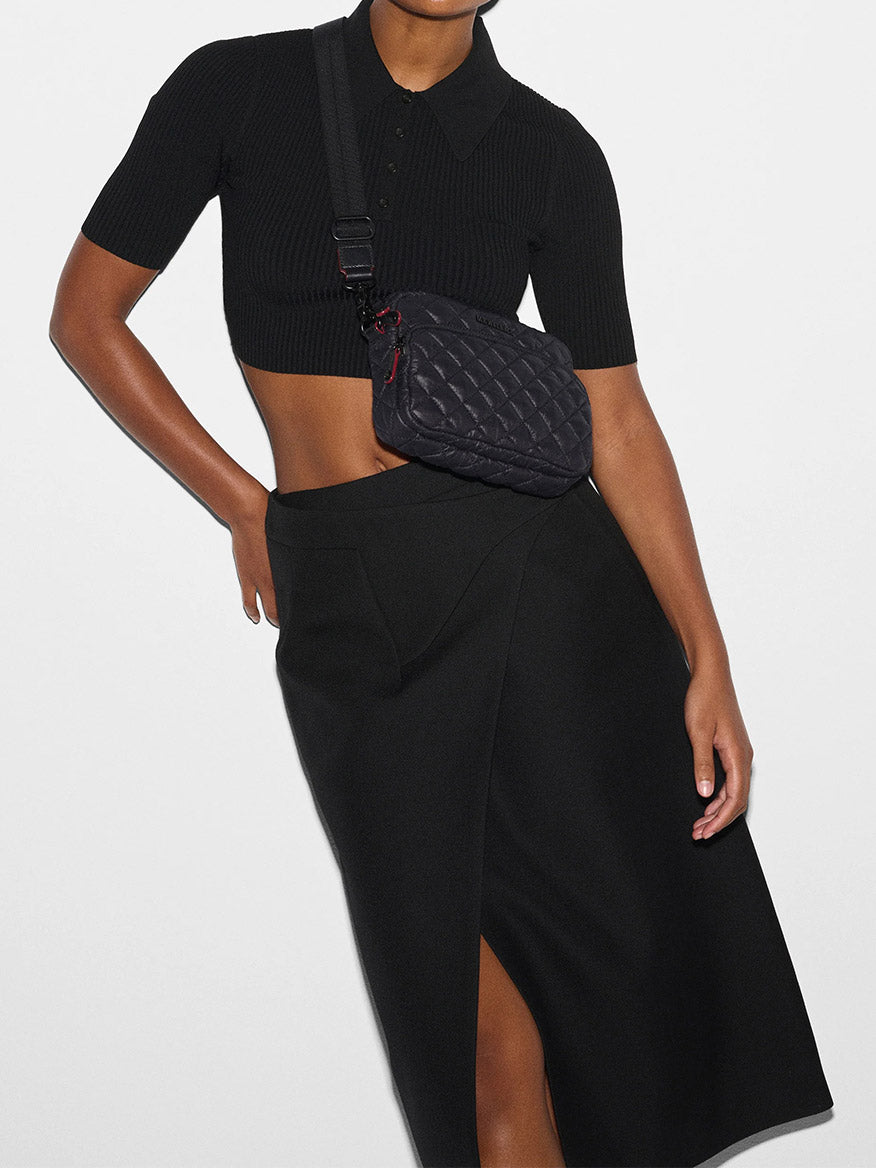 Person wearing a black short-sleeve crop top, a black skirt with a front slit, and an MZ Wallace Small Metro Camera Bag in Black Oxford against a plain white background.