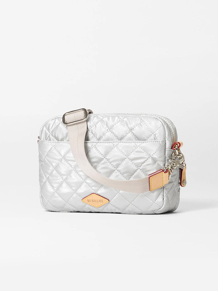 The MZ Wallace Small Metro Camera Bag in Oyster Metallic Oxford features quilted silver nylon, a beige adjustable strap, leather trim, a tan leather patch, and a zippered closure, shown against a plain white background.