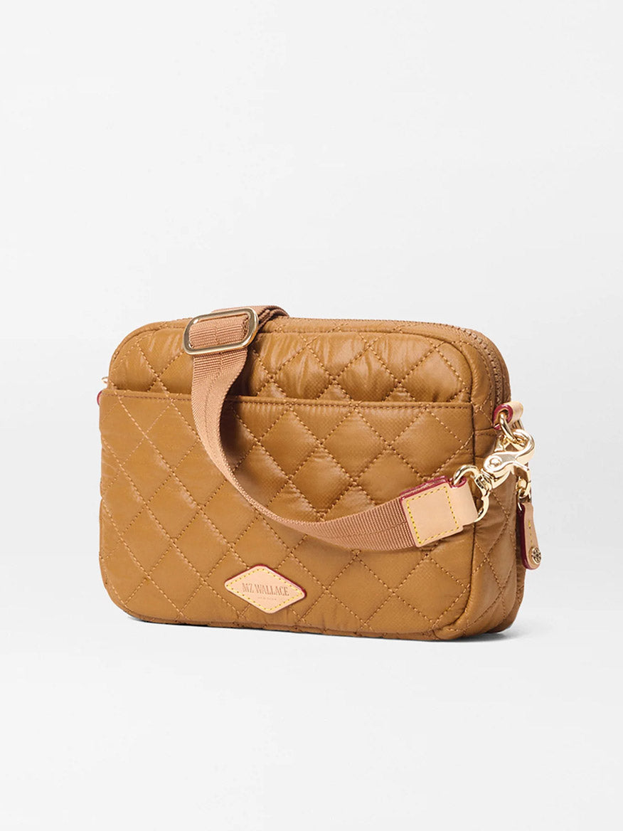 The MZ Wallace Small Metro Camera Bag in Amber Oxford is a compact, rectangular nylon crossbody with quilted design, gold clasp, adjustable strap, and front logo patch, shown against a plain white background.