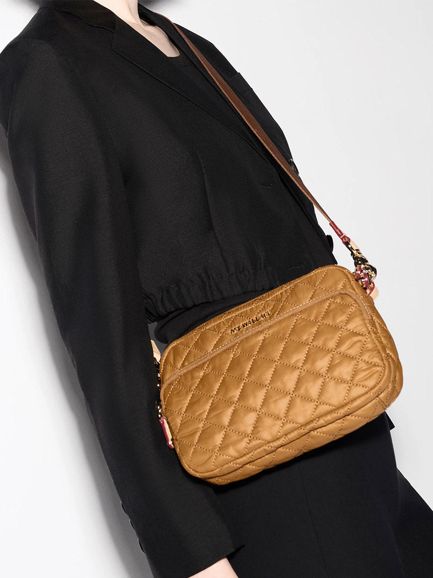 A person in a black outfit carries the MZ Wallace Small Metro Camera Bag in Amber Oxford, a small quilted light brown nylon handbag with a shoulder strap.
