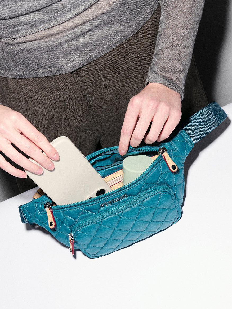 A person places a smartphone into the MZ Wallace Small Metro Sling in Pacific Oxford, a teal quilted waist bag with an adjustable crossbody strap, which already holds other small items.
