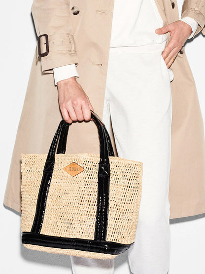 A woman in a trench coat holding a MZ Wallace Small Raffia Tote in Raffia/Black with padded nylon handles.