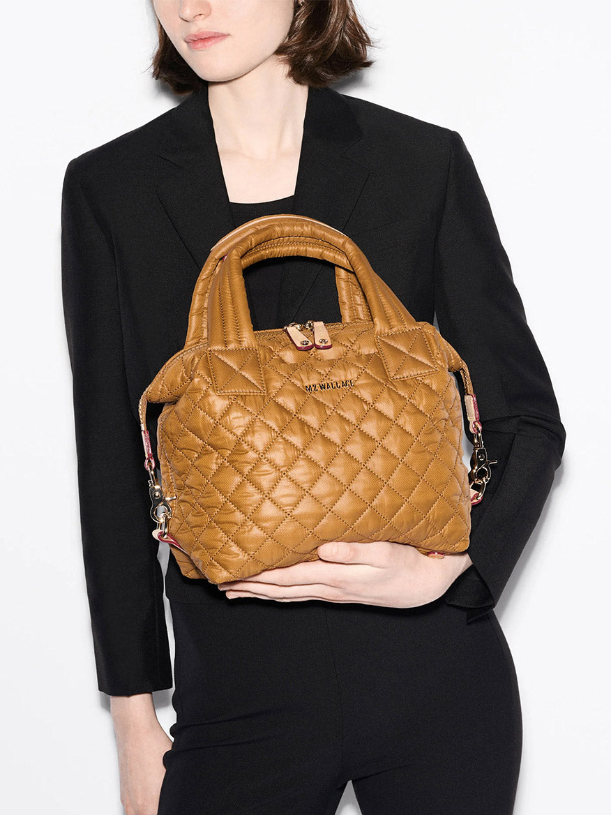 A person in a black blazer holds the MZ Wallace Small Sutton Deluxe in Amber Oxford, a quilted crossbody bag with short handles and a detachable strap.