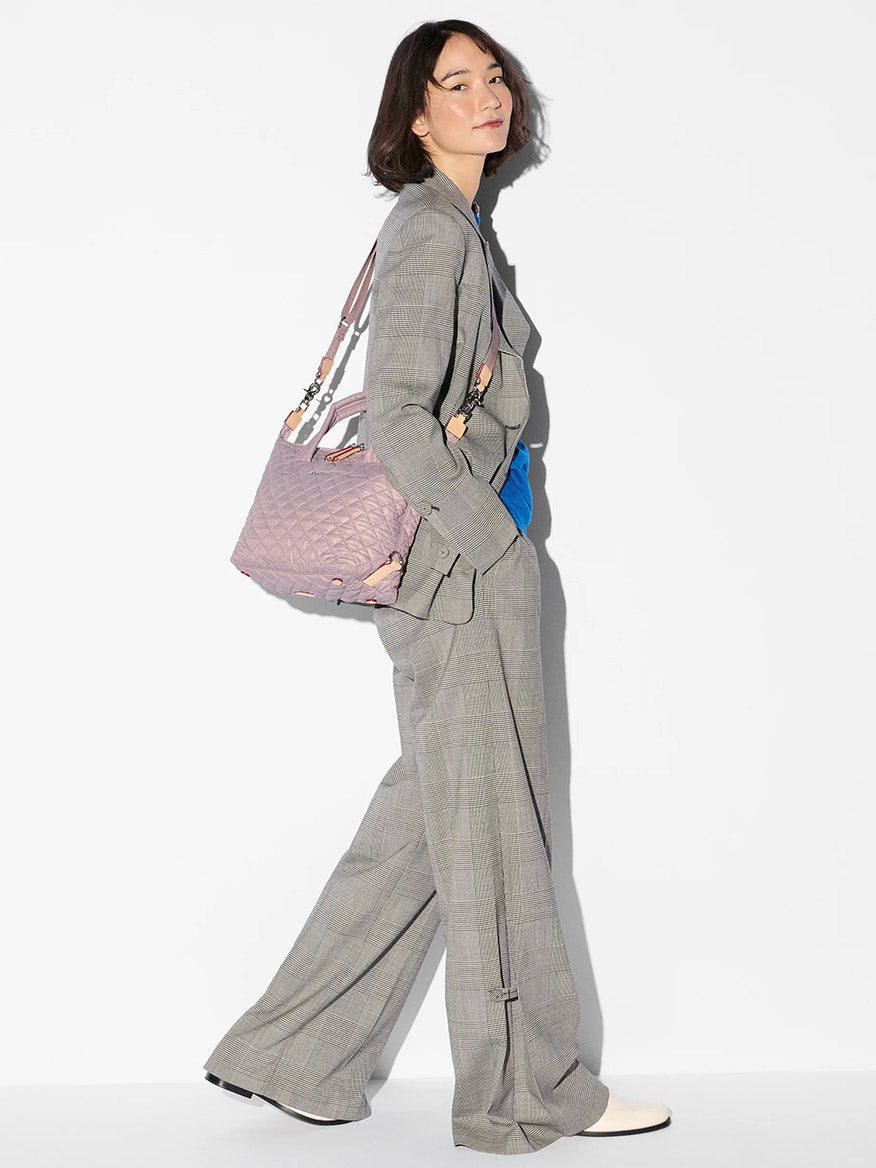 Woman in a gray plaid suit, blue shirt, and white shoes poses sideways, carrying the MZ Wallace Small Sutton Deluxe in Quail Oxford.