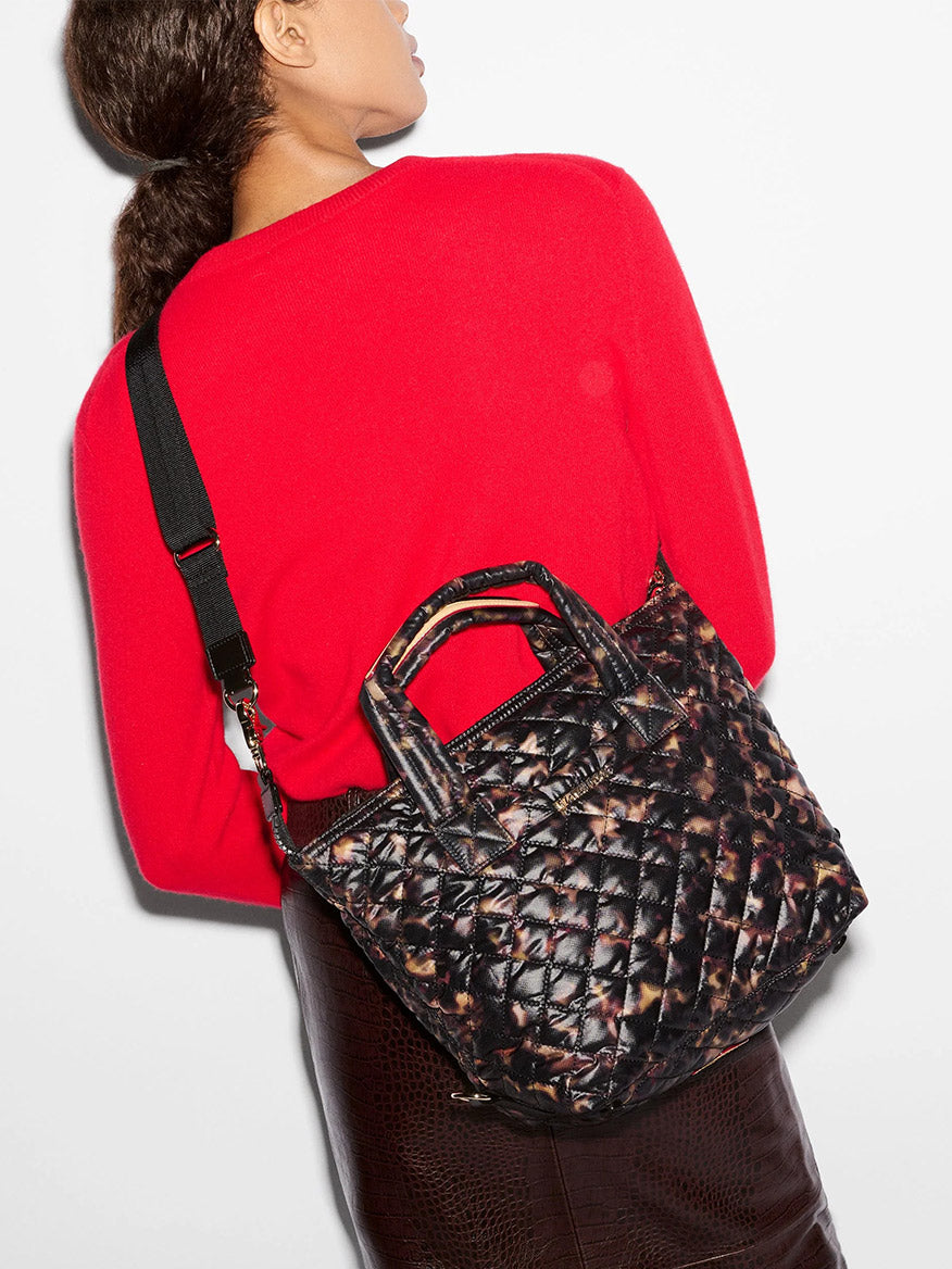 Person wearing a red sweater and brown skirt carrying an MZ Wallace Small Sutton Deluxe in Tortoise Shell Oxford, a small quilted crossbody tote with a brown and black tortoise shell print.