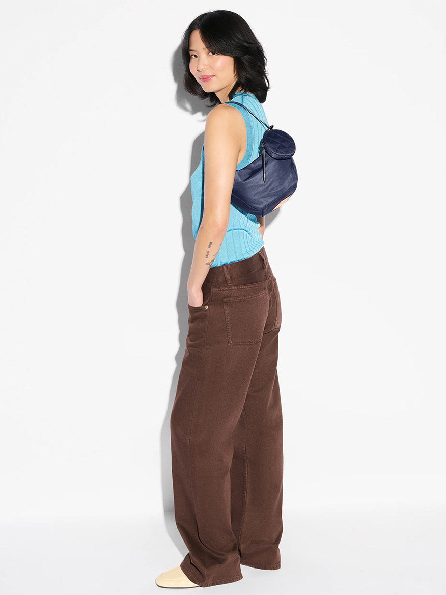 A woman stands sideways in a blue sleeveless top, brown wide-leg pants, cream shoes, and carries the MZ Wallace Small Waverly Hobo in Dawn Bedford—an Italian leather bag with zip top closure—over her shoulder against a white background.
