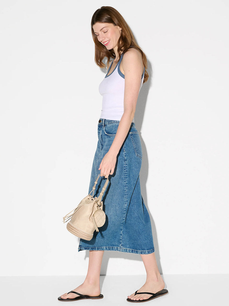 A woman in a white tank top, blue denim midi skirt, and black sandals stands sideways holding the MZ Wallace Waverly Espadrille Bucket Bag in Buff Bedford with Italian leather trim against a plain white background.