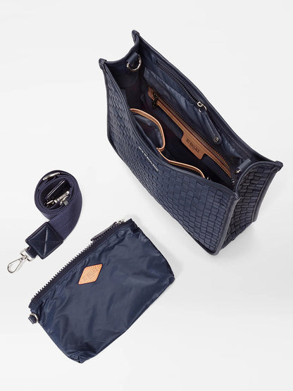 A navy-blue MZ Wallace Woven Box Crossbody in Dawn Oxford open to show the interior, accompanied by a matching zippered pouch and a rolled-up belt with a metallic buckle.