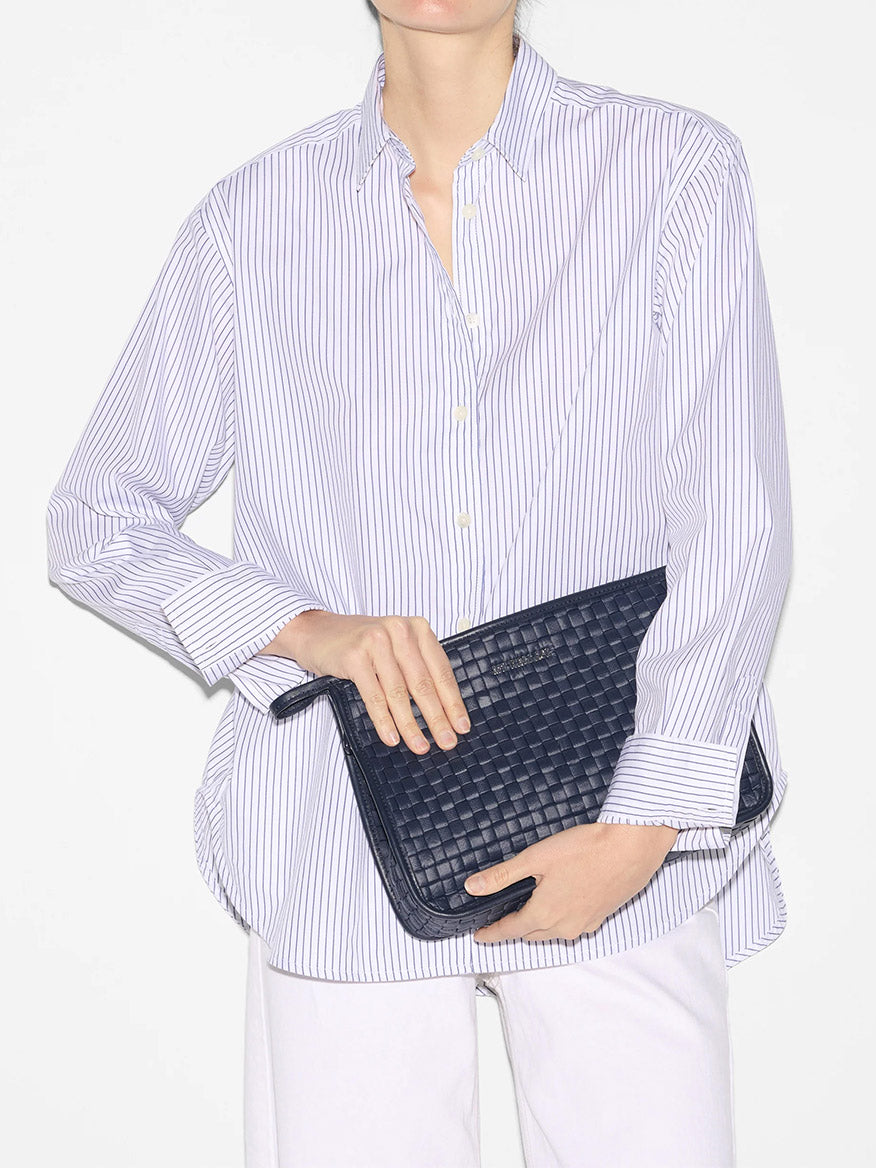 A person wearing a striped blue and white shirt with Italian leather trim and white pants, holding a MZ Wallace Woven Metro Clutch in Dawn Oxford.