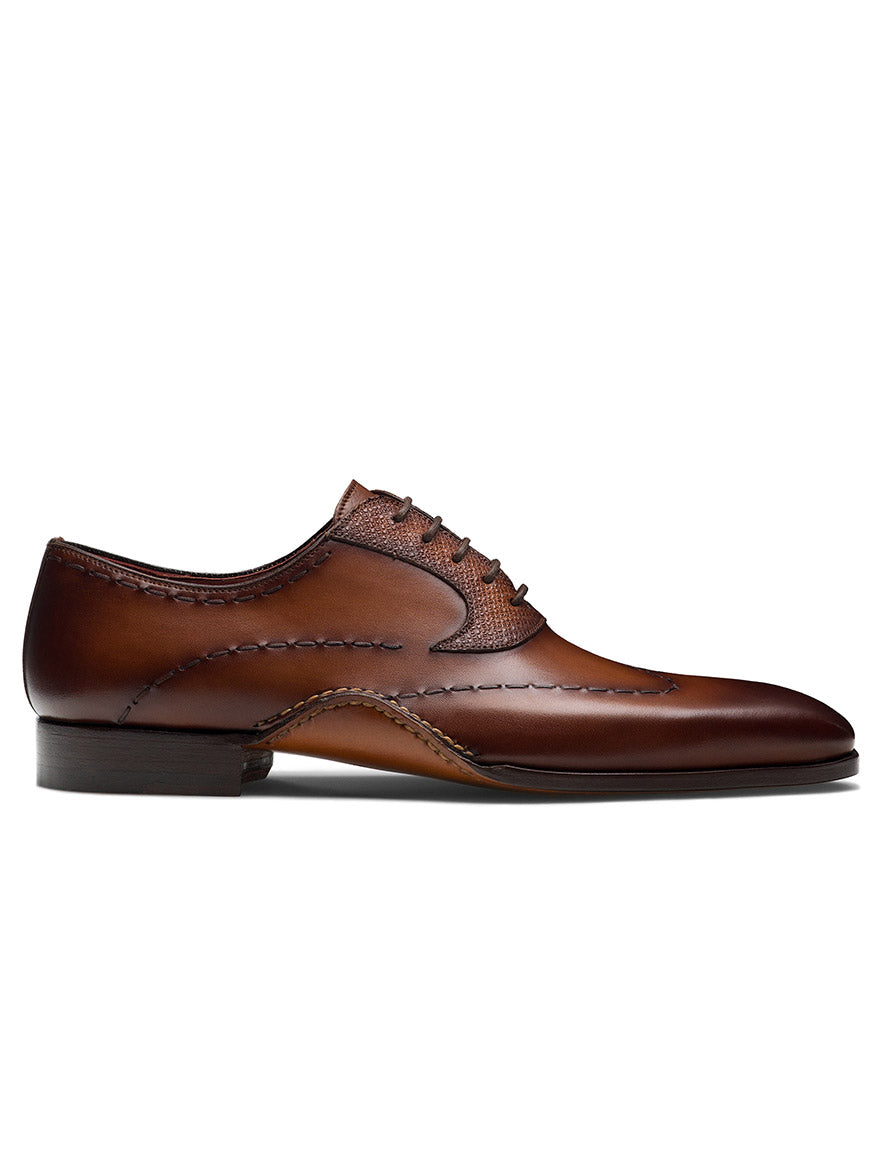 The Magnanni Bowden in Cognac is a brown leather Wingtip Oxford featuring decorative stitching, lace-up closure, and a smooth toe design. Made from premium Grabado leather, this elegant dress shoe embodies timeless style for any formal occasion.