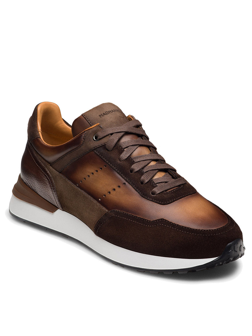 The Magnanni Bravo V in Midbrown Suede/Brown features brown leather and suede, dark brown laces, a white midsole, and black outsole. This luxury men's sneaker is crafted from full-grain leather and is displayed at a slight angle on a plain background.