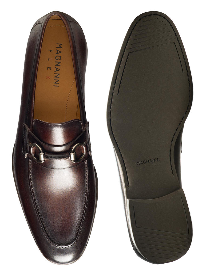 A pair of brown leather slip-on loafers with metallic buckle detail and moccasin toe design, shown from the top and bottom views. The insole reads "Magnanni Diago II in Brown," and the cushioned rubber sole ensures comfort with every step.