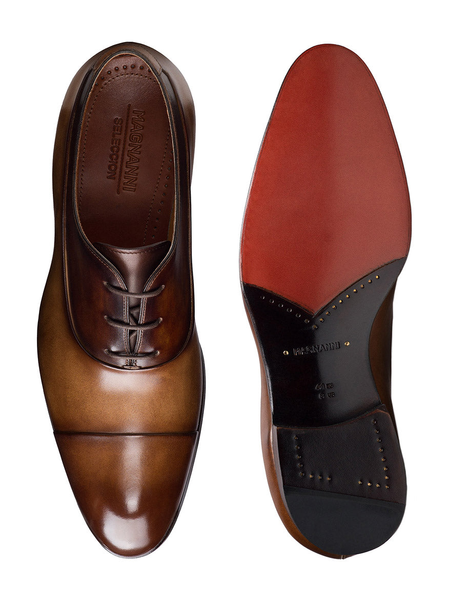 Discover the art of craftsmanship with Magnanni's Duke in Tabaco/Brown leather dress shoes, one beautifully revealing its laces and the other flaunting a chic red sole embossed with the brand name, embodying luxury at every stride.