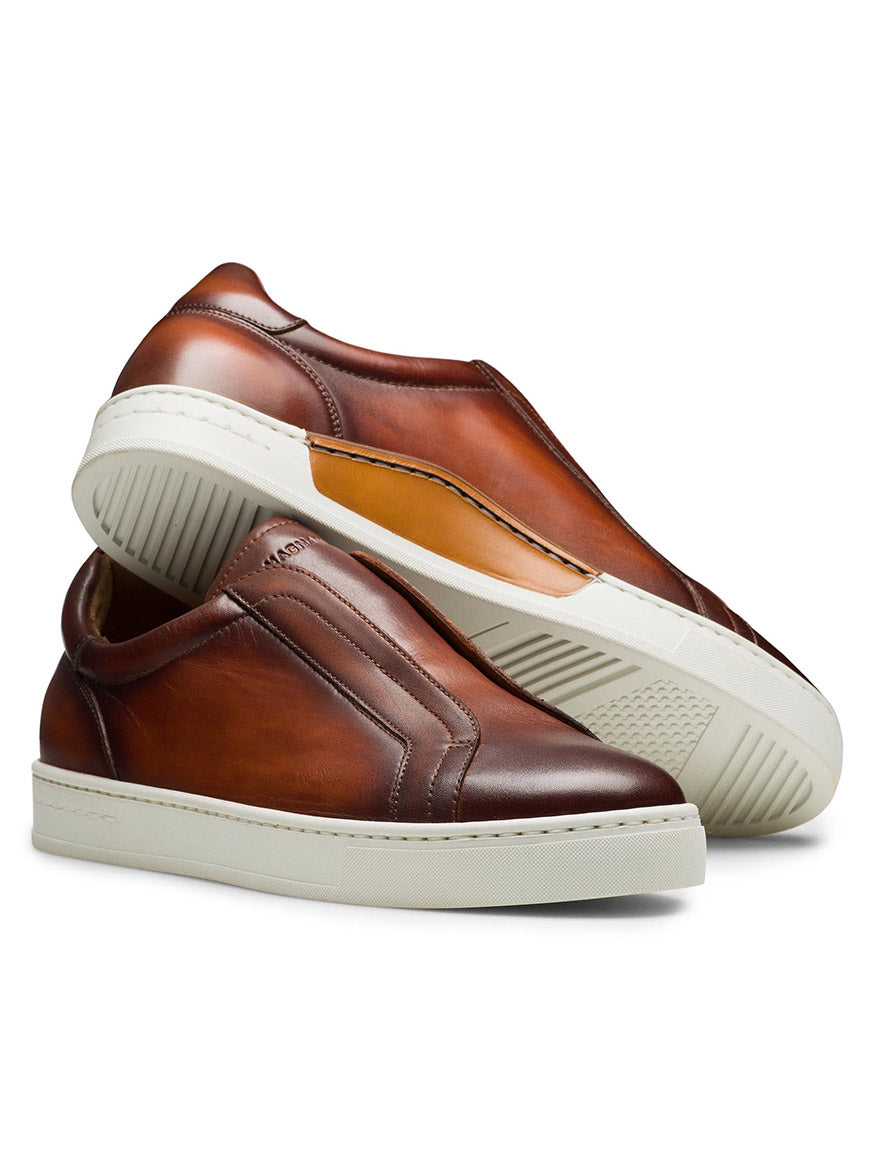 A unique slip-on sneaker, the Magnanni Gasol in Cognac features a pair of brown leather sneakers with a stitched leather insert and elastic gore system for easy wear. One shoe stands upright while the other lies on its side, both boasting white rubber soles.