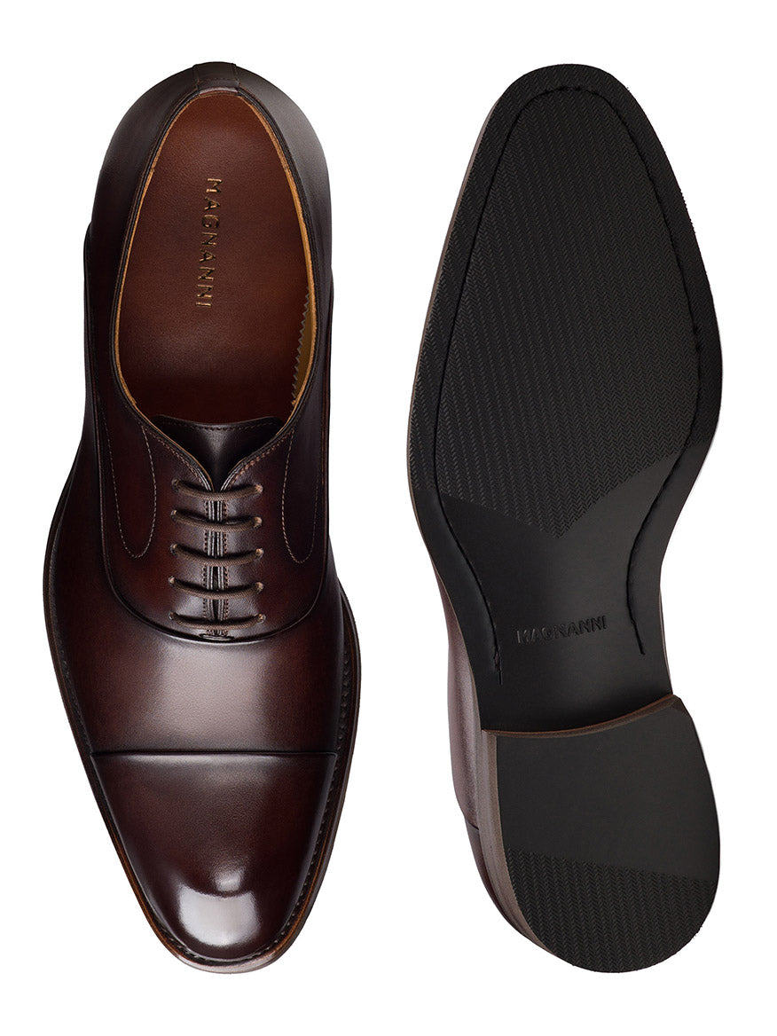 The Magnanni Omega in Midbrown features polished calfskin leather and intricate laces from a top view, alongside a textured sole, embodying timeless elegance as they rest side by side.