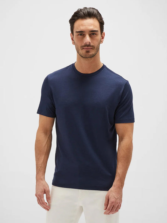 A man stands against a white background wearing a navy blue Maurizio Baldassari Linate Technmerino Short Sleeve T-shirt and white pants, looking straight at the camera with a neutral expression.