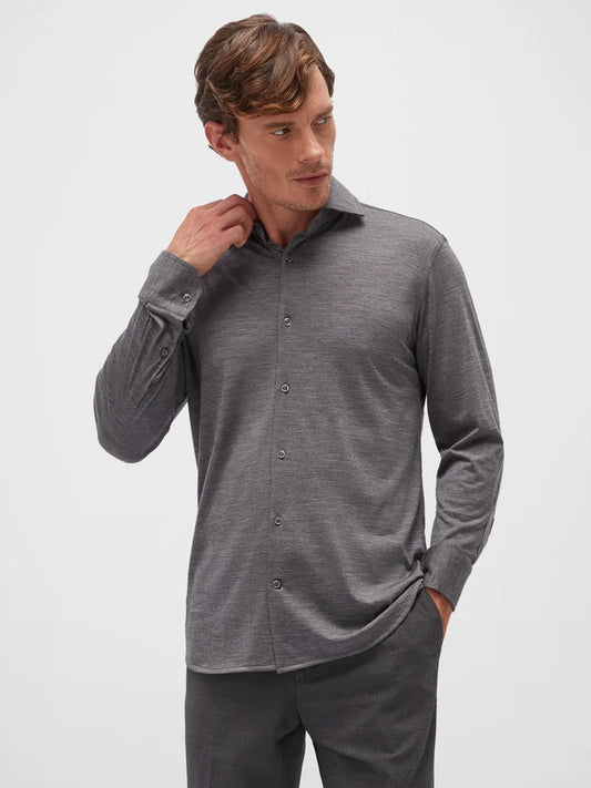 A man models a Dark Grey, long-sleeve Maurizio Baldassari Santini Techmerino Wool shirt and pants, standing against a plain background, with his right hand adjusting his collar.