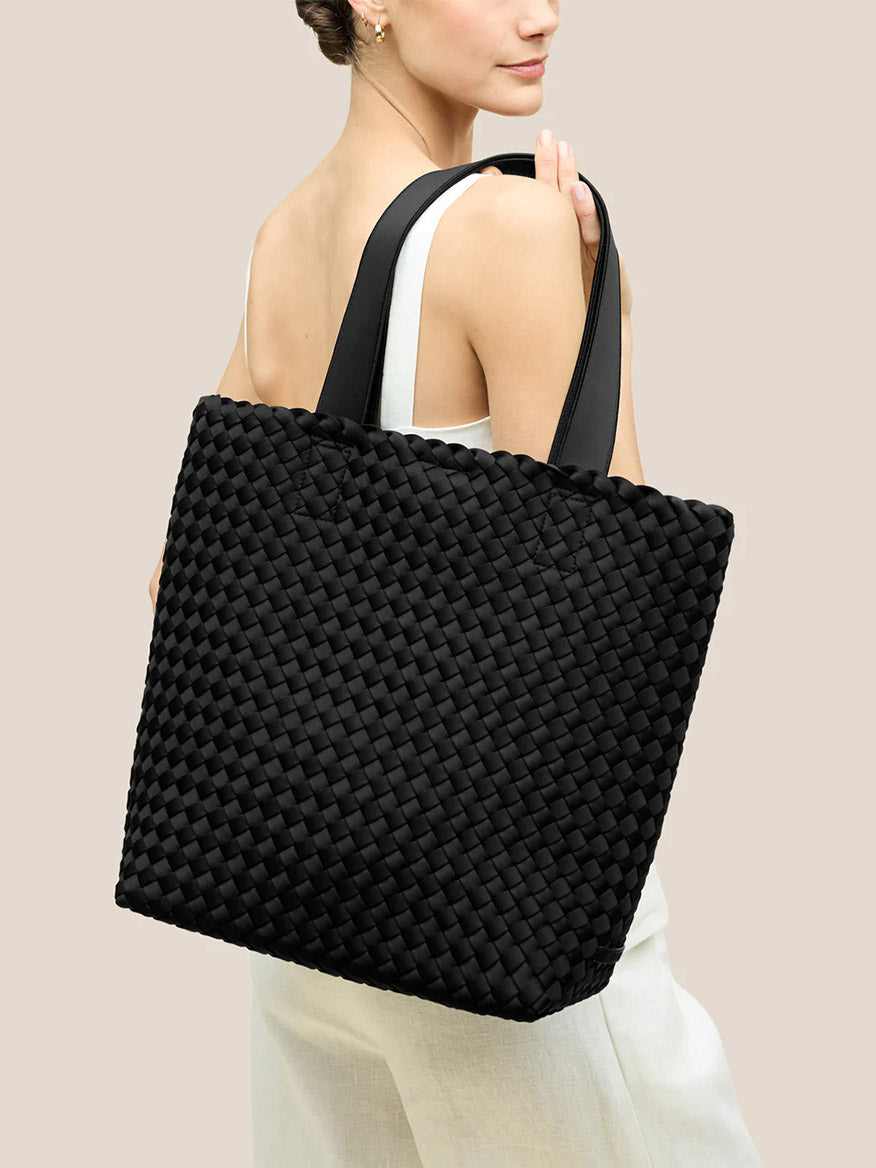 A woman in a white sleeveless dress carries the Naghedi Como Shopper Tote in Onyx over her shoulder, set against a plain beige background.