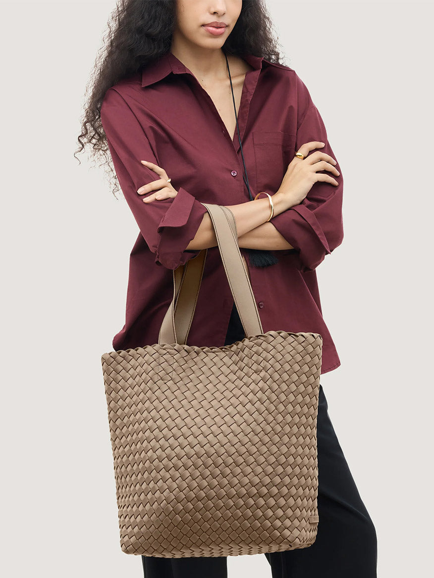 A person wearing a burgundy button-down shirt carries a large, beige Naghedi Como Shopper Tote in Cashmere with wide shoulder straps.