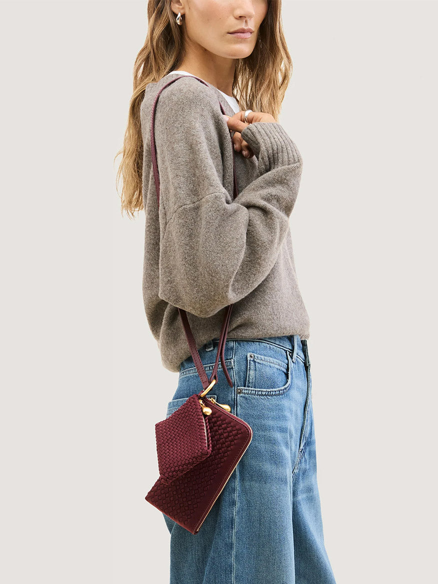 A person wearing a woven bordeaux Naghedi Orly Phone Case with a matching small pouch and an adjustable leather strap.