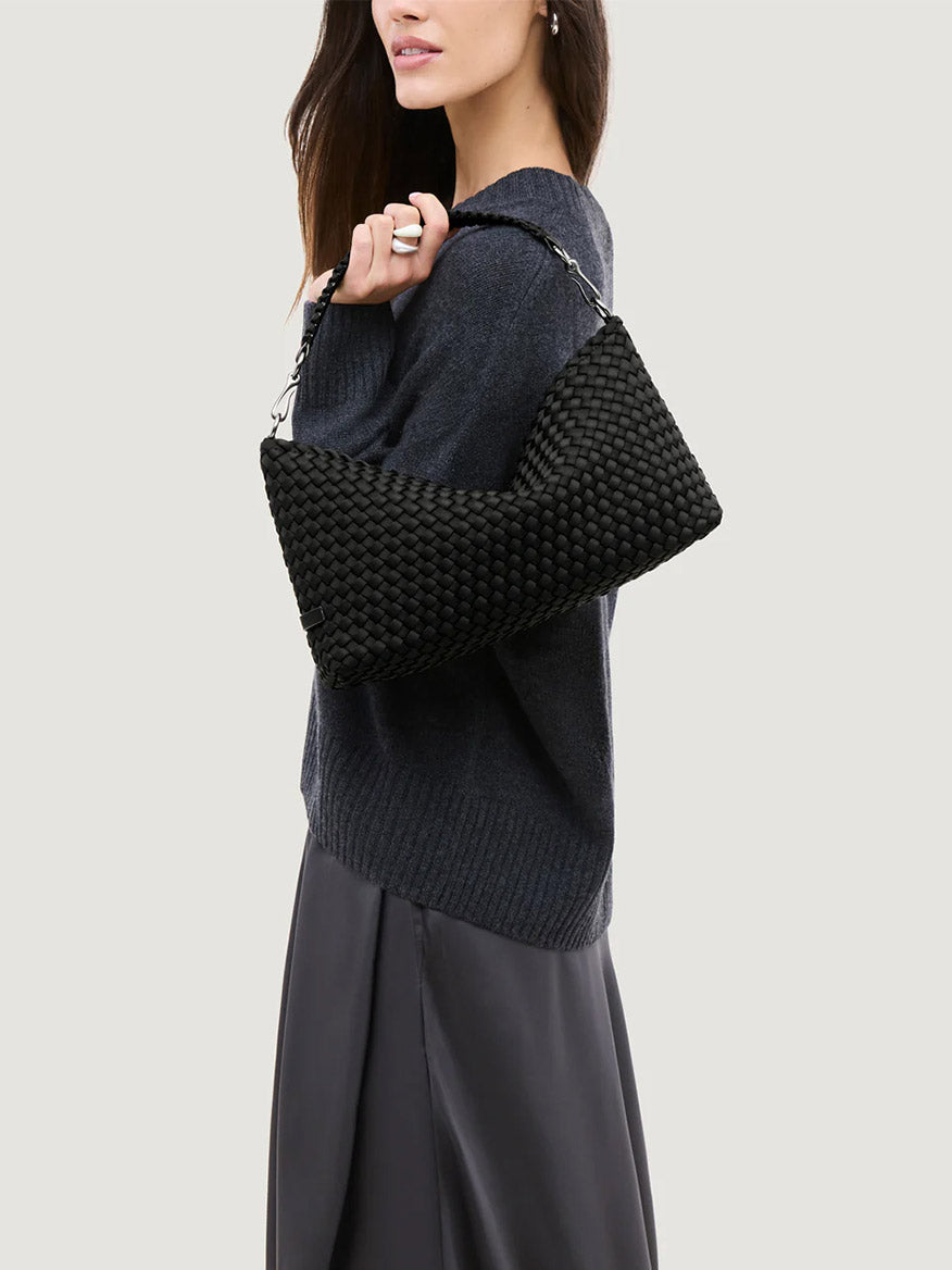 A woman in a dark sweater and skirt holds the Naghedi Porto Small Half-Moon Bag in Onyx under her arm against a plain light background.