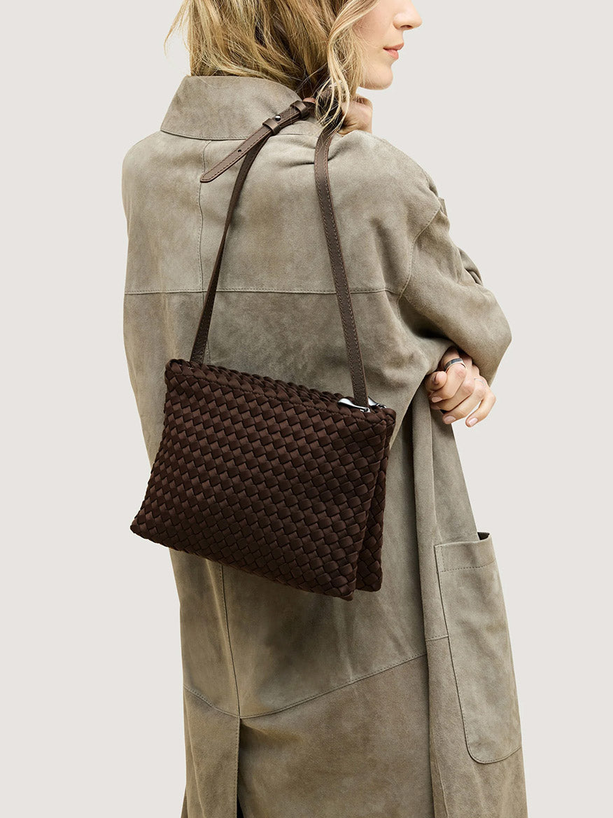 A person wearing a tan coat carries a dark brown handwoven neoprene Naghedi Soho Envelope Crossbody in Chocolate with a thin, adjustable strap.